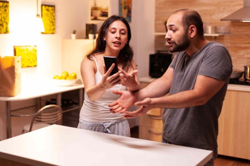 A man and woman stand in a kitchen, the woman showing the man something on a smartphone related to marriage trust, while he gestures towards it, both looking engaged in conversation that may involve consulting a detective agency in Pune.