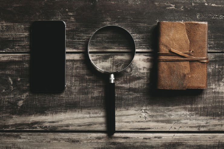 A smartphone, a magnifying glass, and a closed leather journal are neatly arranged on a rustic wooden table at a detective agency. The items convey a theme of investigation or study.